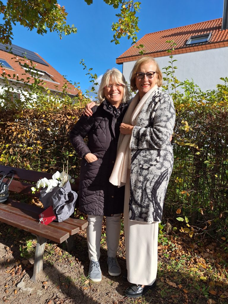 Zwei Frauen stehen im Freien vor einer Hecke, neben einer Bank